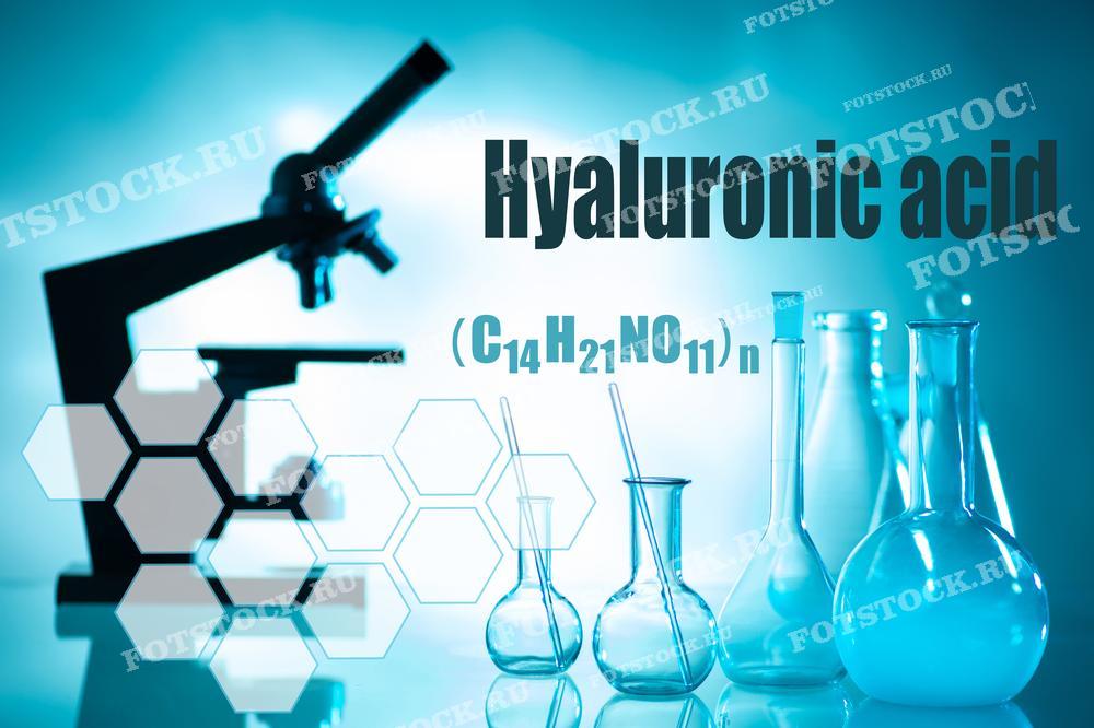 На фотографии изображены надпись "Hyaluronic acid" и химическая формула гиалуроновой кислоты (C14H21NO11)n на фоне лабораторного оборудования, включая микроскоп и различные колбы. Фотография иллюстрирует процесс научных исследований и анализа гиалуроновой кислоты в лаборатории.